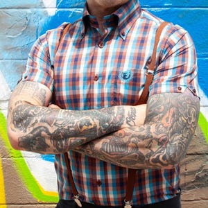 May include: A short-sleeved plaid shirt in shades of blue, orange, and white, featuring a button-down collar and a small emblem on the chest. Brown suspenders are attached to the black pants. The background is a colorful graffiti wall.