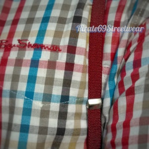 May include: Close-up of a checkered shirt with a red, white, blue, and brown pattern. The shirt has a red strap with a silver clasp. The Ben Sherman logo is embroidered in red on the shirt.