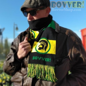 May include: A black, green and yellow striped scarf with a green circle and a black silhouette of a warrior's head. The scarf has the word "BOVVER!" printed on it and the text "BovverShop.com" is visible in the background.