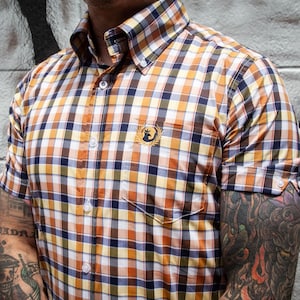 May include: Short-sleeved button-down shirt with a plaid pattern in shades of orange, brown, blue, and white. The shirt features a small pocket and a logo on the chest. The collar is buttoned down.