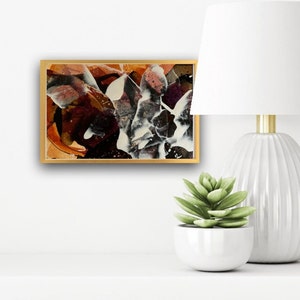 May include: Abstract painting in a gold frame with shades of brown, white, and black. The painting is on a white shelf with a white lamp and a succulent plant in a white pot.
