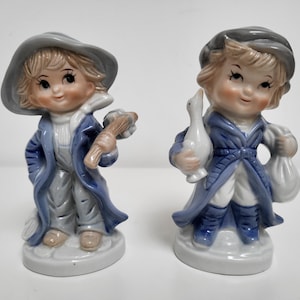 Puede incluir: Dos figuritas de porcelana de niños. La figurita de la izquierda es un niño que lleva un abrigo y un sombrero azules, sosteniendo un palo. La figurita de la derecha es una niña que lleva un abrigo y un sombrero azules, sosteniendo un ganso blanco.