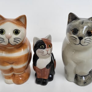 Puede incluir: Tres figuritas de cerámica de gatos. El gato más grande es de color marrón y blanco a rayas. El gato del medio es negro, marrón y blanco. El gato más pequeño es gris y blanco.