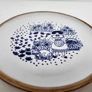 May include: White ceramic plate with a brown rim, featuring a blue illustration of a woman, cat, and flowers. The design includes a woman in a dress, a cat, and a basket of flowers.