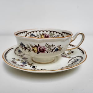 May include: A white teacup and saucer with a floral pattern in shades of pink, yellow, and brown. The cup has a scalloped edge and a gold trim. The saucer has a matching floral pattern and a gold rim.