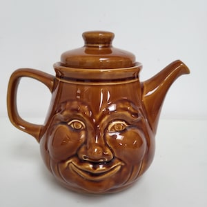 May include: Brown ceramic teapot with a smiling face design. The teapot has a lid, handle, and spout. The face features eyes, a nose, and a wide, cheerful smile. The teapot is a warm, earthy brown colour.