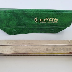 May include: A silver M. Hohner Echo harmonica with a green case. The harmonica is rectangular with the word "ECHO" printed on it. The green case has the words "M. Hohner" and "ECHO" printed on it.