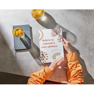 May include: A white card with brown and orange abstract shapes and the text "Believe in yourself & your abilities". The card is being held by a person wearing a yellow shirt.