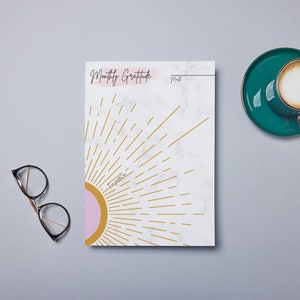 May include: A white marble-patterned notepad with a pink and gold sunburst design. The notepad has the text "Monthly Gratitude" at the top.