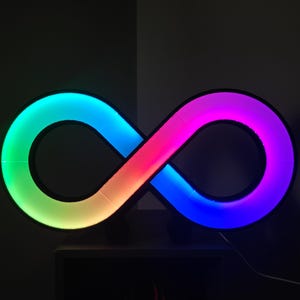 May include: An illuminated infinity symbol with a black border. The light inside the symbol transitions through a spectrum of colors, including green, blue, pink, and purple. The symbol is mounted on a dark wall.