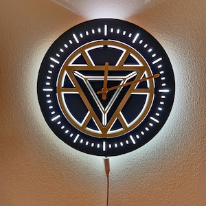 May include: A round, black wall clock with a gold geometric design in the center. The clock has a white illuminated border and gold clock hands. The clock is powered by a cord.