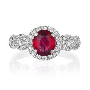 May include: A silver ring with a large red gemstone surrounded by a halo of smaller white gemstones. The ring has two smaller clusters of white gemstones on either side of the center stone.