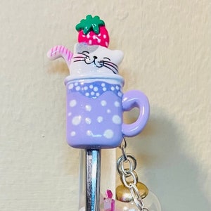 Puede incluir: Un bolígrafo caprichoso con un cuerpo azul marino y una parte superior decorativa con un gato en una taza morada con una fresa en la cabeza. El bolígrafo tiene una sección transparente llena de confeti colorido y un dije de estrella.