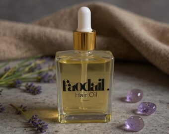 May include: A clear, square glass bottle of hair oil with a gold cap and white dropper. The bottle has the text "faodail. Hair Oil" in black. The background includes lavender sprigs and three purple, round stones.
