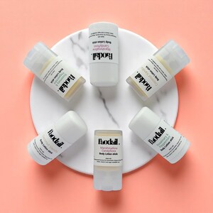 May include: An overhead shot of six white body lotion sticks arranged on a white marble-patterned surface against a pink background. Each stick is labeled with a different scent, including Coconut Vanilla and Marshmallow Candyfloss.