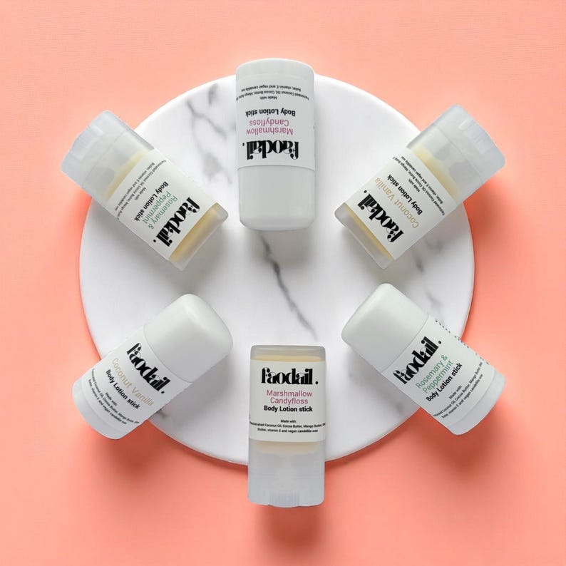 May include: An overhead shot of six white body lotion sticks arranged on a white marble-patterned surface against a pink background. Each stick is labeled with a different scent, including Coconut Vanilla and Marshmallow Candyfloss.