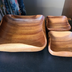 May include: Set of four wooden bowls in various sizes. The bowls are made of light brown wood with a natural grain pattern. The bowls are all square shaped with rounded edges.