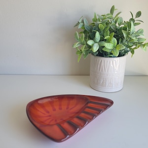 May include: A red ceramic ashtray with a scalloped edge and six slots for cigarettes. The ashtray is on a white surface.