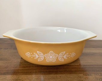 Pode incluir: Um prato de cozedura de cerâmica amarelo com um design floral branco. O prato tem duas alças e é feito de Pyrex.