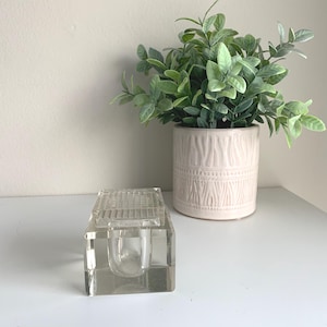 May include: A clear, rectangular glass inkwell with a grid-like top sits on a white surface. A small, round opening is visible inside. A decorative, light pink ceramic pot with a green plant is in the background.