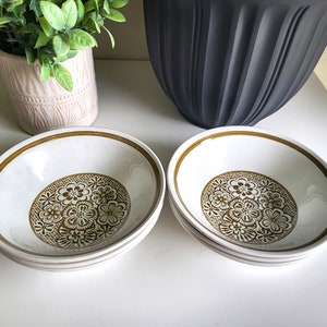 Peut inclure: Quatre bols en céramique blanche avec un motif floral marron au centre. Chaque bol a un bord marron. Les bols sont empilés sur une surface blanche. Un pot rose avec de la verdure est en arrière-plan.