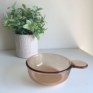 May include: An amber glass bowl with a handle. The bowl is round with a flared rim and a handle on one side. A white ceramic pot with a green plant is in the background.