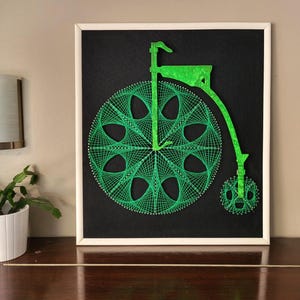 May include: Green string art depicting a vintage bicycle with a large front wheel and a smaller back wheel. The art is framed in a white frame and mounted on a black background.