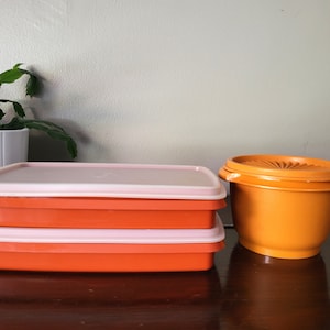 May include: Three orange plastic containers with lids. Two of the containers are stacked on top of each other. The top container has a pink lid. The third container is round and has a lid with a decorative design.