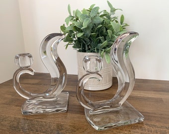 Pode incluir: Dois castiçais de vidro transparente com um design curvo único. Os castiçais estão sobre uma superfície de madeira com uma planta verde em um vaso branco atrás deles.