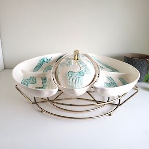 May include: A white ceramic serving dish with a green swirl design. The dish is divided into four sections and has a matching lid. The dish is on a gold metal stand.