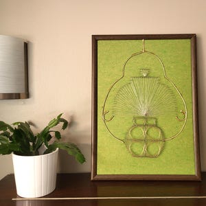 May include: A framed green string art piece depicting a vase with flowers. The vase is made of gold wire and the flowers are white string.