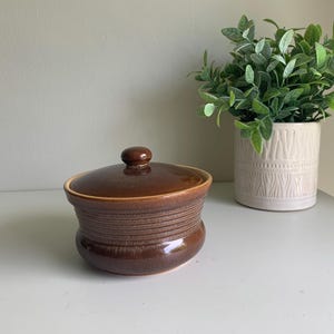 May include: A round, dark brown ceramic lidded jar with a glossy finish and horizontal ribbed detailing around the centre. The lid features a small, rounded knob handle at the top, and the base of the jar has a smooth, slightly wider profile.