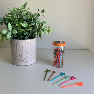 May include: A white ceramic pot with a green plant, a clear container of colourful cocktail sticks, and several sticks scattered on a white surface. The sticks have decorative tops in pink, orange, and green.