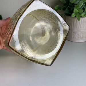 Pode incluir: Um recipiente de vidro de formato quadrado com um tom &acirc;mbar claro e uma base circular lisa. As superf&iacute;cies externas apresentam um padr&atilde;o floral em relevo que cobre os lados, enquanto a parte inferior permanece clara e lisa.