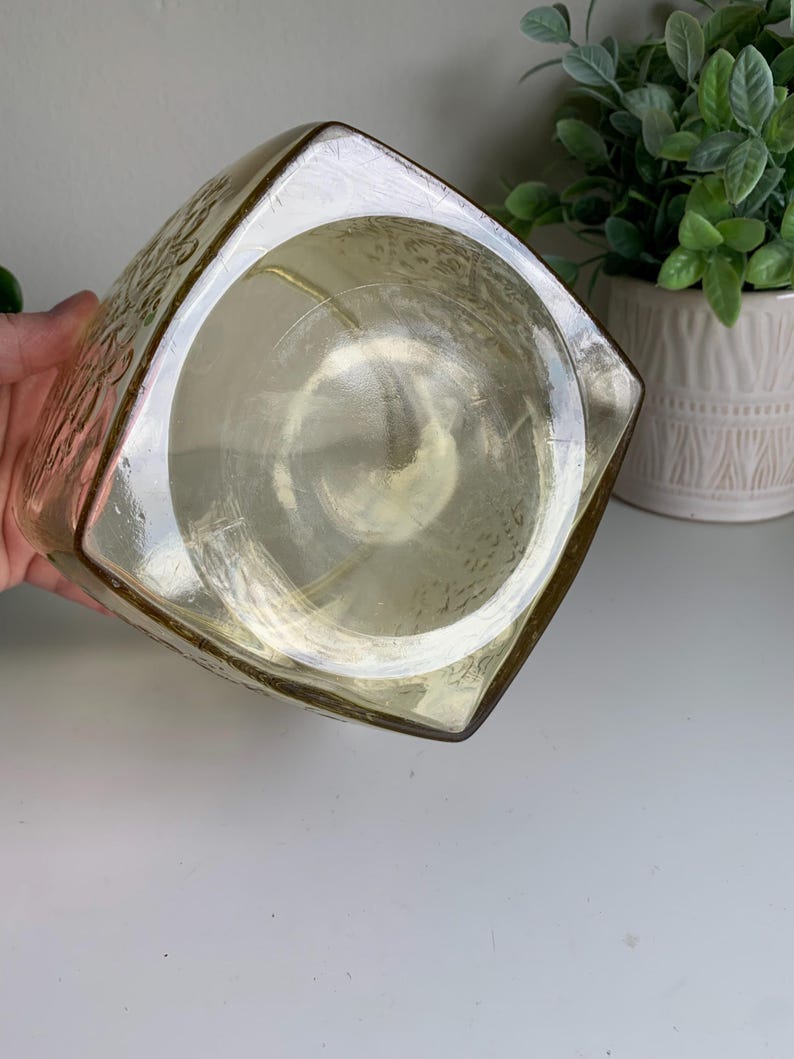 Pode incluir: Um recipiente de vidro de formato quadrado com um tom &acirc;mbar claro e uma base circular lisa. As superf&iacute;cies externas apresentam um padr&atilde;o floral em relevo que cobre os lados, enquanto a parte inferior permanece clara e lisa.