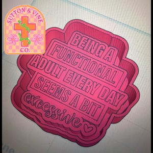 May include: A pink cookie cutter with the text "BEING A FUNCTIONAL ADULT EVERY DAY SEEMS A BIT excessive" in a layered design. The upper left corner features a logo with a cross, flowers, and the text "SUTTON & VINE CO."