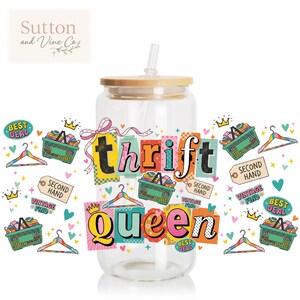 Op de afbeelding: Een doorzichtige glazen beker met een bamboe deksel en rietje. De beker is versierd met de woorden "thrift queen" in kleurrijke blokletters, omringd door afbeeldingen van winkelmanden, kleerhangers en labels.
