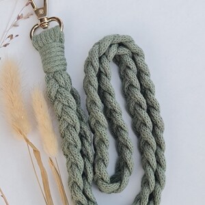Könnte beinhalten: Ein geflochtenes Lanyard in Salbeigrün mit einem goldenen Metallverschluss. Das Lanyard besteht aus einem weichen, gewebten Stoff.