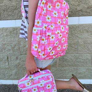 Pode incluir: Uma mochila rosa brilhante e uma lancheira a condizer com um padrão de grelha branca sobreposto por margaridas brancas e caras sorridentes amarelas. Ambos os artigos têm fechos e rebordos brancos com um design floral e de caras.