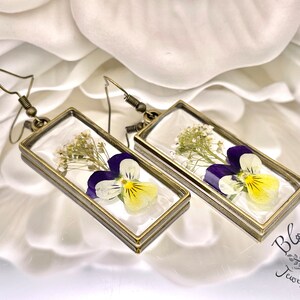 May include: A pair of rectangular earrings with a pressed flower design. Each earring features a purple and yellow pansy and small white flowers encased in a clear resin within a bronze-colored frame. The earrings have bronze-colored ear wires.