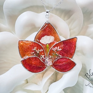 May include: A silver chain necklace with a pendant shaped like a lotus flower. The pendant is made of clear resin with a red and orange glitter background and white pressed flowers inside. The pendant is approximately 2 inches in diameter.