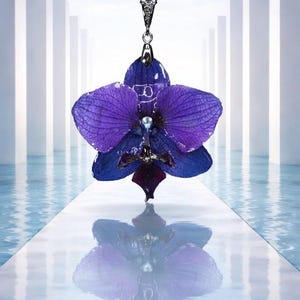 May include: A silver necklace pendant featuring a vibrant purple and blue orchid flower. The pendant is suspended from a delicate chain and reflects in the water below. The orchid has a pearl-like center.