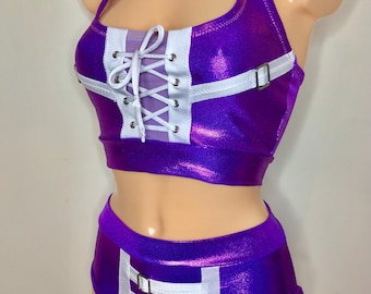 Puede incluir: Un conjunto de dos piezas compuesto por un top corto tipo halter de color morado metalizado y pantalones cortos de talle alto a juego. El top presenta un frente con cordones blancos, ojales plateados y tiras blancas con hebillas plateadas.