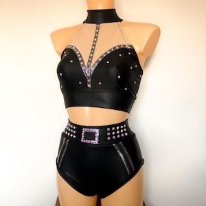Puede incluir: Un traje de baile negro de dos piezas con un top halter y pantalones cortos de cintura alta. El top tiene un panel de malla transparente y est&aacute; adornado con pedrer&iacute;a. Los pantalones cortos tienen un detalle de hebilla plateada y tambi&eacute;n est&aacute;n adornados con pedrer&iacute;a.