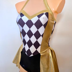 May include: A gold and black leotard with a harlequin pattern on the bodice. The halter-style top features a sheer neckline and gold trim. The leotard has a black bottom and a gold skirt.