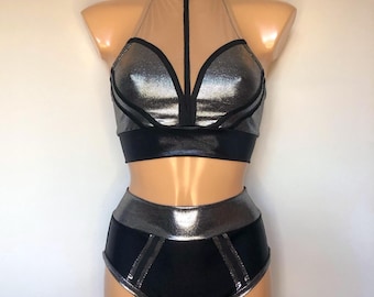 Puede incluir: Un conjunto de baile plateado y negro con un top halter y pantalones cortos de cintura alta. El top tiene un panel de malla transparente y los pantalones cortos tienen una cintura plateada.