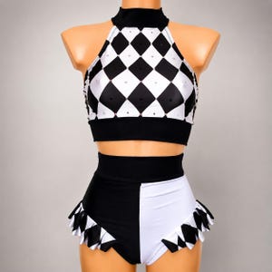 Könnte beinhalten: Ein zweiteiliges Tanzkostüm in Schwarz und Weiß. Das Neckholder-Top hat ein Rautenmuster mit Strasssteinen, ein schwarzes Band und einen hohen Ausschnitt. Die hoch taillierten Shorts sind geteilt, eine Seite schwarz und die andere weiß, und mit einem Rüschen-Rautenmuster verziert.
