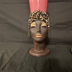 May include: A black mannequin head with a leopard print turban and a pink ceramic vase on top. The mannequin has gold eyeshadow and gold hoop earrings.
