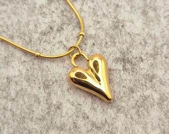 Può includere: Una collana con catena dorata con un ciondolo a forma di cuore. Il ciondolo è un cuore pieno e liscio con una parte inferiore leggermente appuntita.