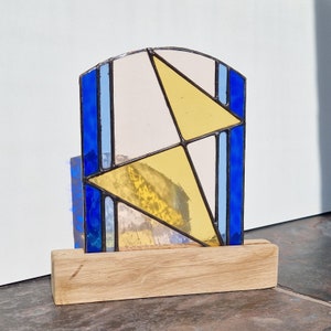 Op de afbeelding: Een geometrisch glas-in-loodpaneel met blauwe, gele en witte vormen, geplaatst op een houten voet. Het abstracte ontwerp toont hoekige vormen en een gebogen bovenkant. De houten voet in natuurlijke kleur contrasteert met het gekleurde glas.